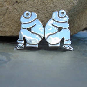 May include: A silver pendant featuring two figures sitting back to back, with their heads bowed. The figures are stylized and have a smooth, polished finish.