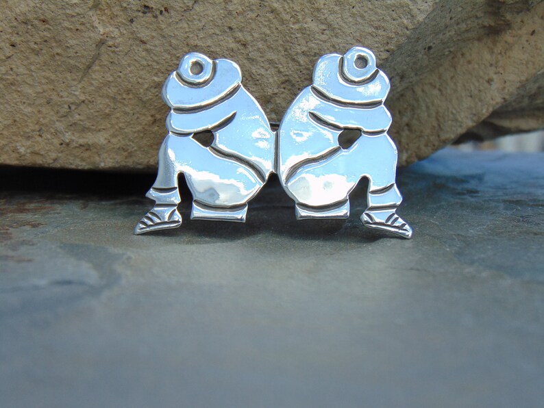 May include: A silver pendant featuring two figures sitting back to back, with their heads bowed. The figures are stylized and have a smooth, polished finish.