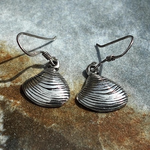 May include: A pair of silver-toned drop earrings featuring a textured, ridged seashell design. Each earring is suspended from a curved metal hook, showcasing a detailed, realistic clam shell shape with horizontal grooves and a polished metallic finish.