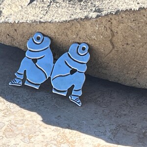 May include: Two silver-toned metal figurines of people sitting with their arms crossed. The figurines are facing each other and are positioned on a light gray surface.