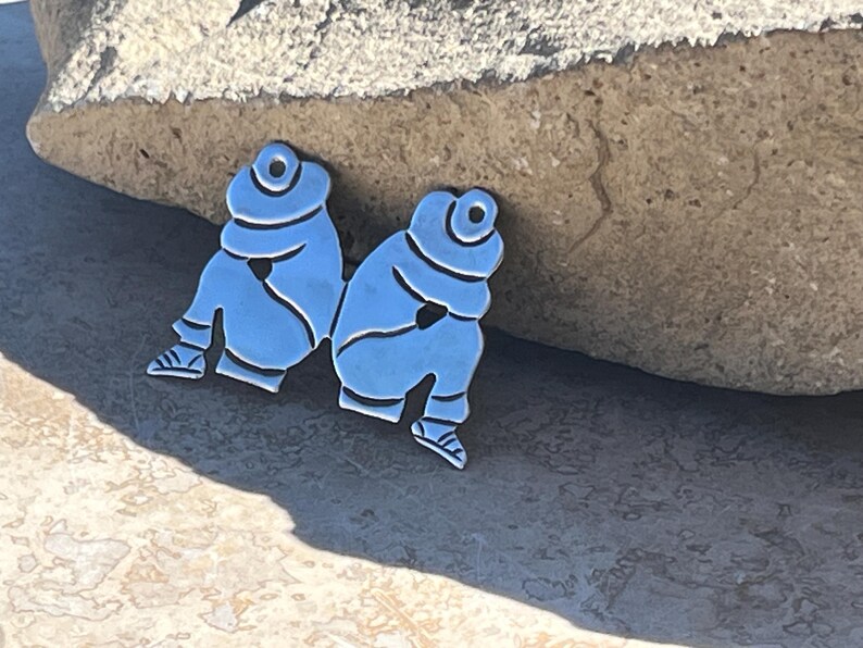 May include: Two silver-toned metal figurines of people sitting with their arms crossed. The figurines are facing each other and are positioned on a light gray surface.