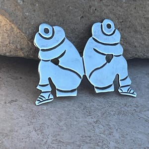 May include: Two silver-toned metal figures, each with a rounded head and arms crossed over their chest, are joined together at the waist. The figures are sitting with their legs bent and their feet pointing outward.