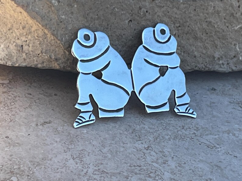 May include: Two silver-toned metal figures, each with a rounded head and arms crossed over their chest, are joined together at the waist. The figures are sitting with their legs bent and their feet pointing outward.
