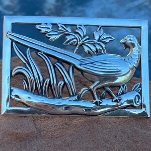 May include: A silver-toned rectangular brooch featuring a detailed pheasant design. The bird is perched on a branch amidst foliage, all within a rectangular frame. The brooch is likely made of metal and has a polished finish, suitable for a jacket or scarf.