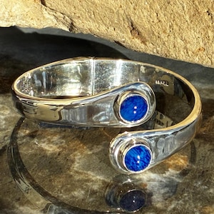 Peut inclure: Un bracelet manchette ouvert en métal argenté poli, orné de deux pierres cabochons circulaires bleu profond à chaque extrémité. La surface métallique est lisse et réfléchissante, avec une charnière visible sur le côté de l'anneau.