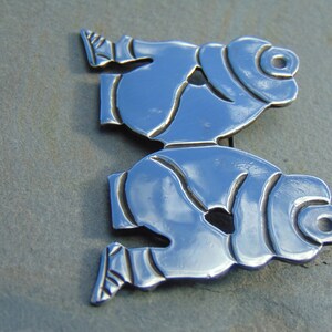 May include: A silver brooch depicting two stylized figures, possibly representing people or animals, with a smooth, reflective surface.