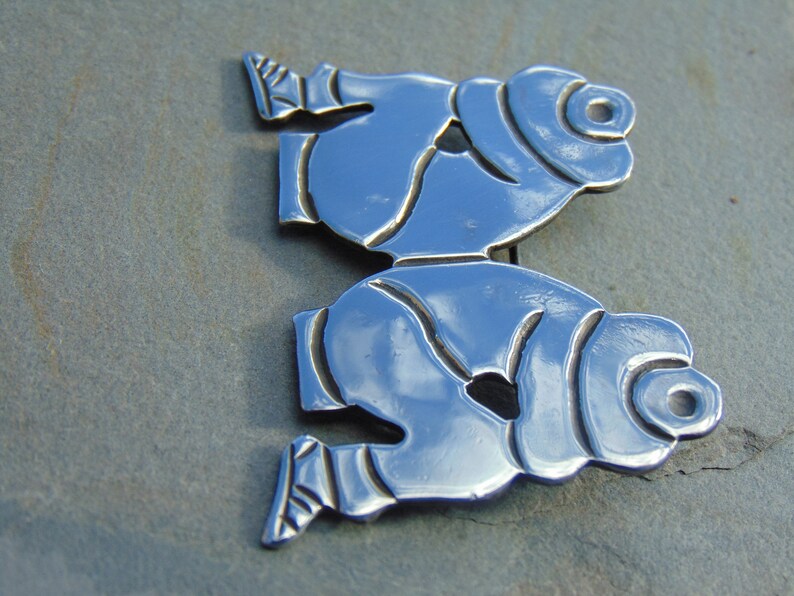 May include: A silver brooch depicting two stylized figures, possibly representing people or animals, with a smooth, reflective surface.
