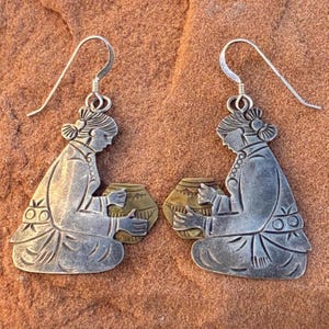 Puede incluir: Pendientes de plata con una figura sentada sosteniendo una olla. La figura tiene detalles grabados en la ropa y el pelo, y la olla es de color dorado. Los pendientes tienen cierre de gancho.
