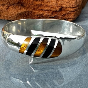 May include: A polished silver cuff bracelet featuring a central inlay of alternating diagonal stripes of golden-brown tiger's eye gemstone and smooth black onyx. The metal band has a wide, curved design with a reflective finish and a tapered overlap.