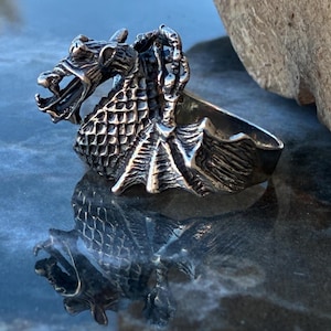 Op de afbeelding: Een gedetailleerde zilveren drakenring. De draak heeft een open mond, schubben en ingewikkelde vleugels. De ring wordt getoond op een reflecterend oppervlak, dat de kenmerken van de draak en zijn reflectie laat zien.