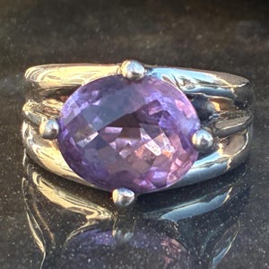 May include: A silver ring featuring a large, faceted, round purple gemstone. The ring has a wide band with a textured design and small silver accents around the gemstone. The ring is displayed on a reflective surface.