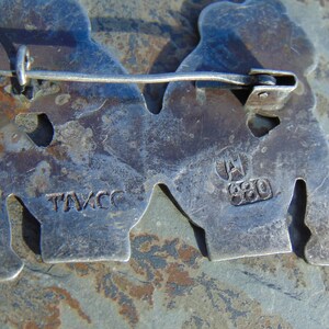 May include: Close-up of the back of a silver pin with two figures. The pin has a clasp on the back and is marked "TAVCC" and "A 980".