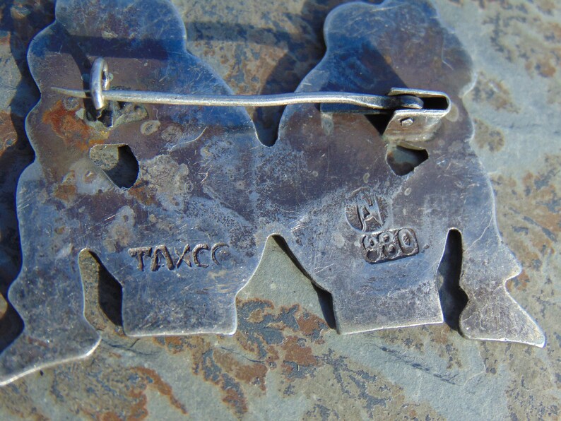 May include: Close-up of the back of a silver pin with two figures. The pin has a clasp on the back and is marked "TAVCC" and "A 980".