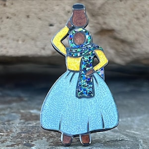 May include: A blue and yellow enamel pin depicting a woman in a blue dress and a yellow shirt carrying a brown pot on her head. The woman is wearing a blue and green patterned scarf.