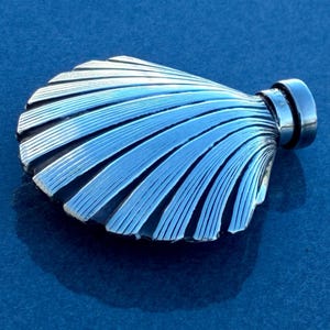 May include: A silver-coloured, seashell-shaped flask with a ridged design. The flask has a small, cylindrical cap on one end. The shell design features alternating ridges and grooves, creating a textured surface. The flask is displayed on a blue surface.