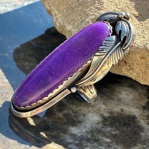 May include: A long, oval-shaped purple stone ring set in a silver band. The setting features intricate metalwork resembling layered feathers or leaves along the side of the stone, with a scalloped edge framing the vibrant, deep purple cabochon.