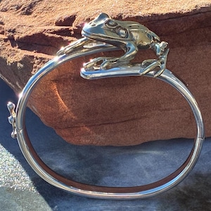 Könnte beinhalten: Silberfarbenes Armreif-Armband mit detailliertem Frosch-Design. Der Frosch sitzt auf einem gebogenen Steg, das Armband umschließt das Handgelenk. Das Armband hat einen Klappverschluss. Innendurchmesser ca. 6 cm.