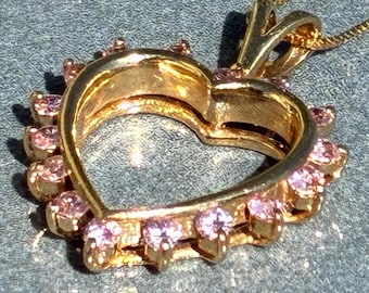 Könnte beinhalten: Eine goldfarbene Herz-Anhänger-Halskette mit einem Kranz aus kleinen rosa Edelsteinen. Der herzförmige Anhänger ist in der Mitte offen und hängt an einer zarten Goldkette. Die Edelsteine sind gleichmäßig angeordnet.