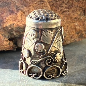 May include: A detailed, antique silver thimble with intricate embossed patterns. The thimble features a perforated top and ornate designs, including floral and geometric shapes. The metal has a tarnished appearance, suggesting age and use.