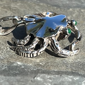 Puede incluir: Un collar con un colgante de plata en forma de cangrejo con ojos de piedras preciosas verdes. El cangrejo está detallado con garras texturizadas y una concha.