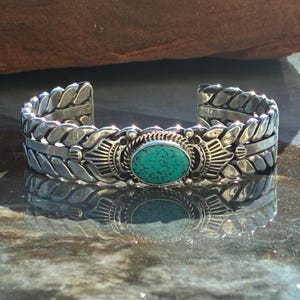 May include: A silver cuff bracelet with an oval turquoise stone centrepiece. The bracelet features a detailed leaf and geometric design. The turquoise stone is a vibrant green-blue colour, set in a decorative silver bezel. The bracelet is displayed on a reflective surface.