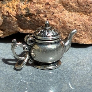 May include: A small, silver-colored teapot charm with intricate details. The charm features a lid, spout, and handle, and is likely made of metal. The teapot is set against a neutral background.