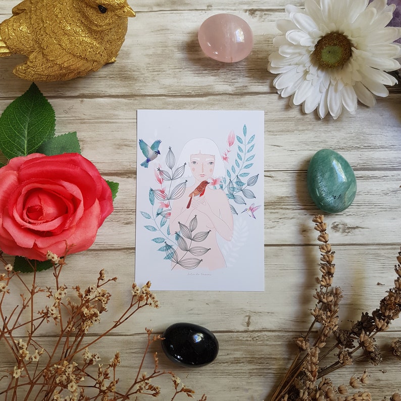 May include: A white card with a drawing of a woman with white hair and a red bird in her hands. The woman is surrounded by green leaves and flowers. The card has the text 'Little Bird Charm' at the bottom.