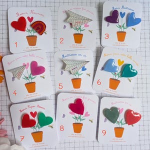 Peut inclure: Neuf cartes de présentation blanches avec des broches en forme de cœurs et d'avions en papier. Chaque carte illustre une plante en pot avec du texte français comme Merci Nounou ou Maitresse en or. Les broches sont des cœurs brillants ou des avions.