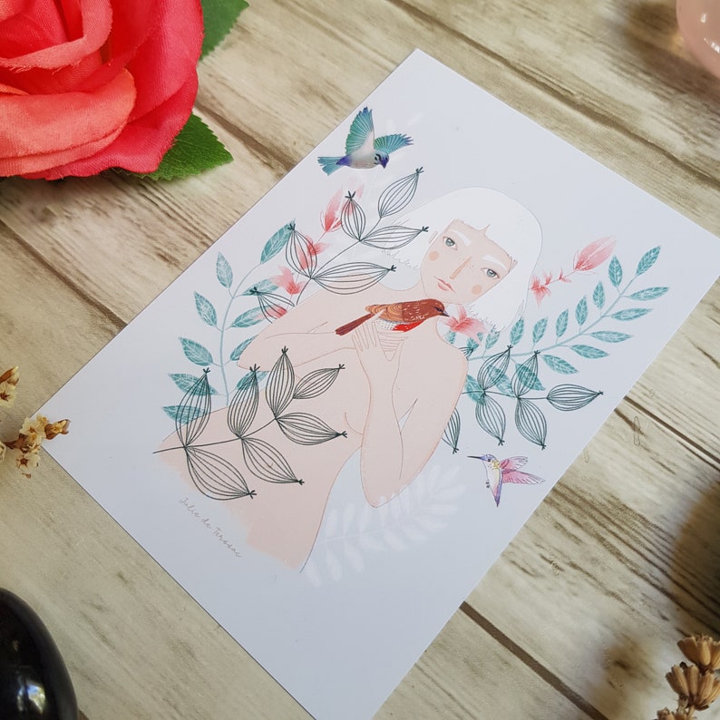 May include: A white card with a woman with white hair and a red bird in her hand. The woman is surrounded by green leaves and pink flowers. The text on the card reads 'Jule de Provence'.