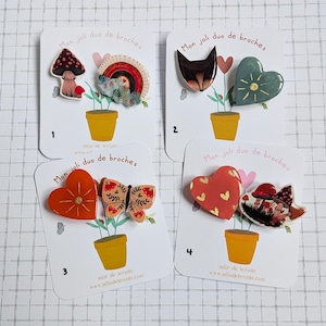 May include: Four sets of decorative brooches on cards. Each card features two brooches with a plant pot design. Brooches include a mushroom, rainbow, cat, heart, butterfly, and gnome. The text "Mon joli duo de broches" is visible.