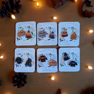 Peut inclure: Six ensembles de broches émaillées sur cartes. Chaque carte présente deux épingles sur le thème de l'hiver, notamment des pulls et des maisons en pain d'épices. Les cartes comportent du texte en français et sont disposées sur une surface en bois.