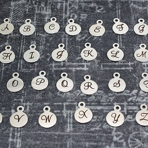 K&ouml;nnte beinhalten: Ein Satz von 26 silberfarbenen Metall-Anh&auml;ngern, jeder mit einem anderen Buchstaben des Alphabets. Die Anh&auml;nger sind rund und haben eine kleine Schlaufe oben zum Anbringen an einer Halskette oder einem Armband.