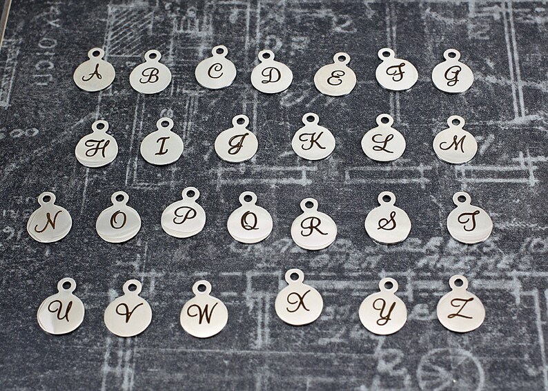 K&ouml;nnte beinhalten: Ein Satz von 26 silberfarbenen Metall-Anh&auml;ngern, jeder mit einem anderen Buchstaben des Alphabets. Die Anh&auml;nger sind rund und haben eine kleine Schlaufe oben zum Anbringen an einer Halskette oder einem Armband.