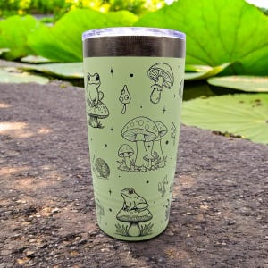 May include: A light green stainless steel tumbler with a black lid. The tumbler features a whimsical design of frogs and mushrooms in black outline.