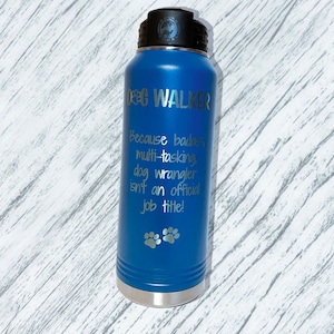 Puede incluir: Una botella de agua aislada azul con tapa negra. La botella tiene las palabras "DOG WALKER" y una cita humor&iacute;stica sobre los adiestradores de perros. Dos huellas de patas est&aacute;n en la parte inferior de la botella.