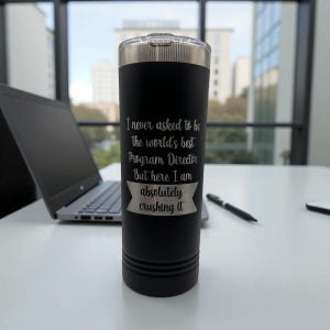 Puede incluir: Un vaso t&eacute;rmico negro con tapa plateada. El vaso tiene grabado el texto "I never asked to be the world's best Program Director But here I am absolutely crushing it". Un port&aacute;til y un bol&iacute;grafo est&aacute;n en el fondo.