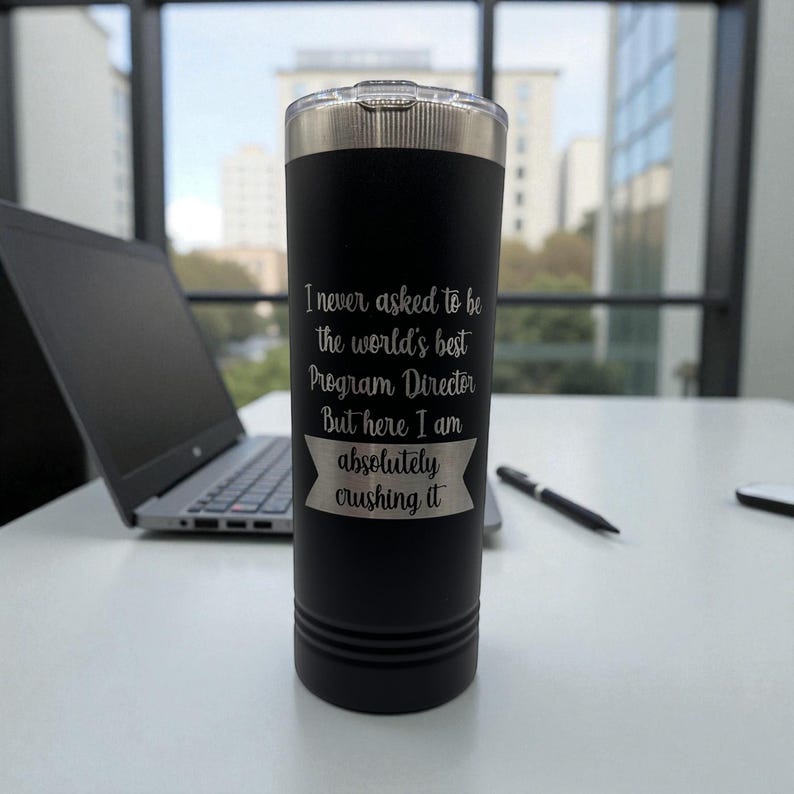 Puede incluir: Un vaso t&eacute;rmico negro con tapa plateada. El vaso tiene grabado el texto "I never asked to be the world's best Program Director But here I am absolutely crushing it". Un port&aacute;til y un bol&iacute;grafo est&aacute;n en el fondo.