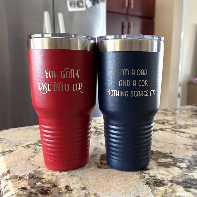 Puede incluir: Dos vasos aislados. Uno es rojo con el texto "You Gotta Ease Into This". El otro es azul marino con el texto "I'm a Dad and a Cop. Nothing Scares Me". Ambos tienen bordes y tapas plateadas.