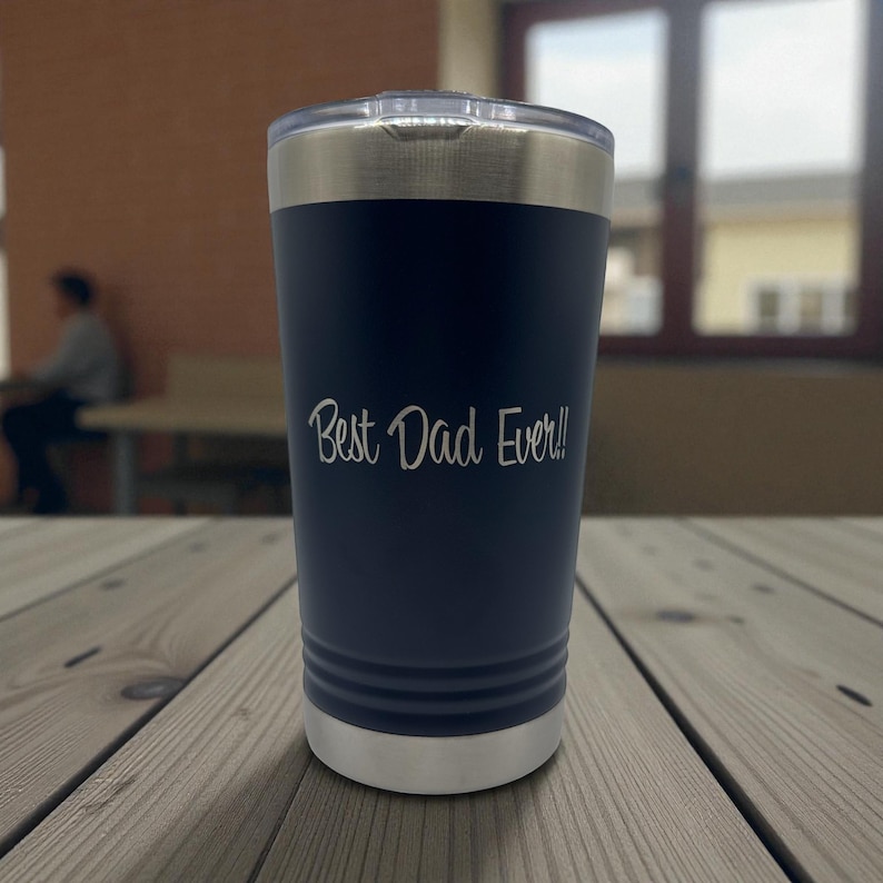 Puede incluir: Un vaso t&eacute;rmico azul marino con borde y tapa plateados. El texto "Best Dad Ever!!" est&aacute; grabado en blanco. El vaso cil&iacute;ndrico est&aacute; sobre una superficie de madera.