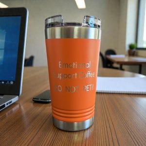 Puede incluir: Un vaso t&eacute;rmico naranja con borde y tapa plateados. El vaso tiene el texto grabado "Emotional Support Coffee DO NOT PET!!". Est&aacute; sobre una mesa de madera, con un port&aacute;til y un tel&eacute;fono en el fondo.