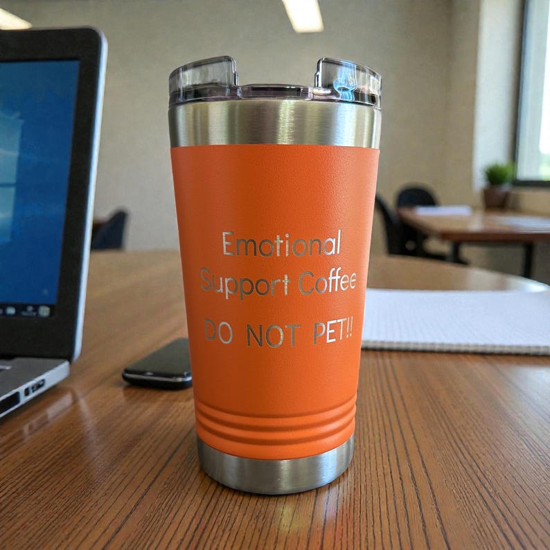 Puede incluir: Un vaso t&eacute;rmico naranja con borde y tapa plateados. El vaso tiene el texto grabado "Emotional Support Coffee DO NOT PET!!". Est&aacute; sobre una mesa de madera, con un port&aacute;til y un tel&eacute;fono en el fondo.