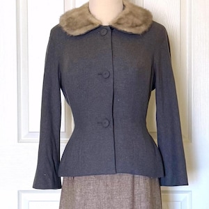 May include: A vintage gray wool suit featuring a jacket with a fur collar and a matching knee-length skirt. The jacket has a fitted waist and button closure. The skirt is a neutral brown color.