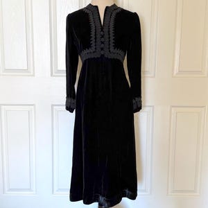 May include: A black velvet dress with long sleeves and a midi-length skirt. The dress features intricate dark gray embroidery around the neckline, bodice, and cuffs. The dress is displayed on a mannequin against a white background.