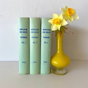 May include: Three green hardcover books titled "Montcalm and Wolfe" by "Parkman" with volumes 1, 2, and 3. A yellow glass vase with yellow daffodils sits in front of the books.