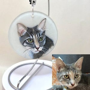 May include: A hand-painted glass ornament featuring a portrait of a gray tabby cat with green eyes. The cat is looking directly at the viewer.
