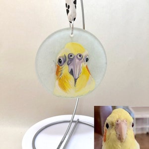 May include: A hand-painted glass ornament featuring a yellow cockatiel with orange and white markings. The ornament is round and has a white background. It is hanging from a silver stand.
