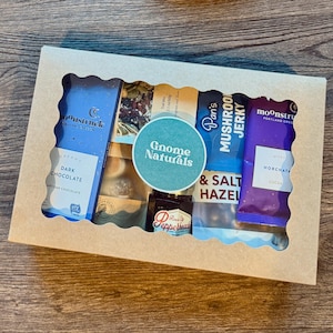 Peut inclure: Un coffret cadeau avec une fenêtre transparente, rempli de diverses collations. Comprend une barre de chocolat noir, un mélange montagnard, du jerky aux champignons et une barre de chocolat horchata. Le coffret a un autocollant "Gnome Naturals".
