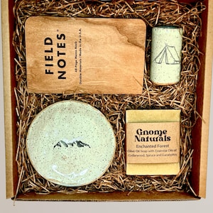 May include: A gift box filled with a small ceramic dish with a mountain design, a wooden notepad with the text "FIELD NOTES", a small ceramic mug with a tent design, and a bar of soap with the text "Gnome Naturals Enchanted Forest Olive Oil Soap with Essential Oils of Cedarwood, Spruce and Eucalyptus".