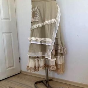 Puede incluir: Falda envolvente beige con múltiples capas de volantes y ribete de encaje. La falda presenta un parche decorativo con detalles de encaje y un cierre de hebilla. La falda se exhibe en un soporte metálico.