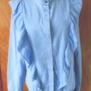May include: Light blue blouse with a high collar and long sleeves. The blouse features ruffled accents along the shoulders and down the front. The fabric appears to be a lightweight material, suitable for warmer weather.